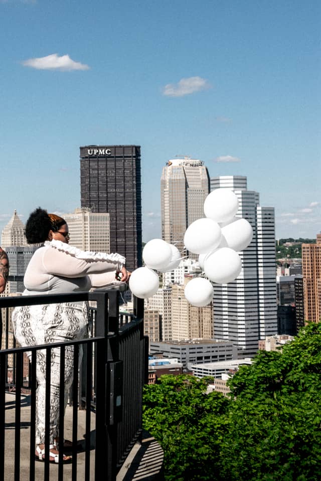 A lady is just about to cut a strand of white balloons tied to a railing with the Pittsburgh skyline in the background