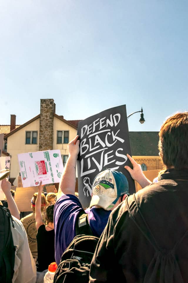 A man is holding a sign that says Defend Black Lives along with a group of others also holding protest signs