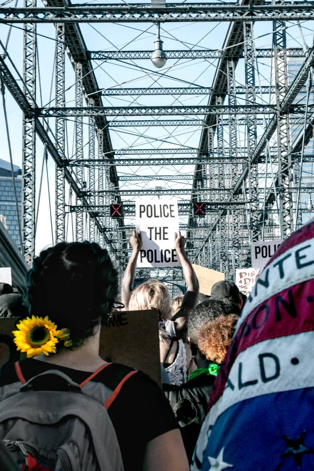 Someone holds a sign that says Police the Police in the middle of a Pittsburgh Black Lives Matter protest. To the left is a woman with a sunflower in her hair, and the right is a flag with the names of those who have died in recent months since the BLM protests started.