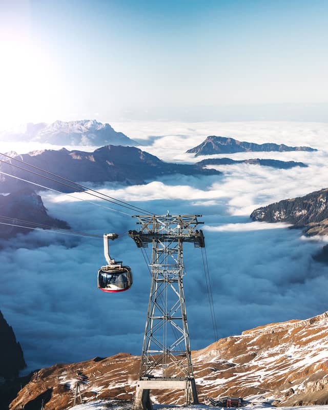 A gandola is shown descending a snow-covered mountain above a large body of clouds and mountain tops