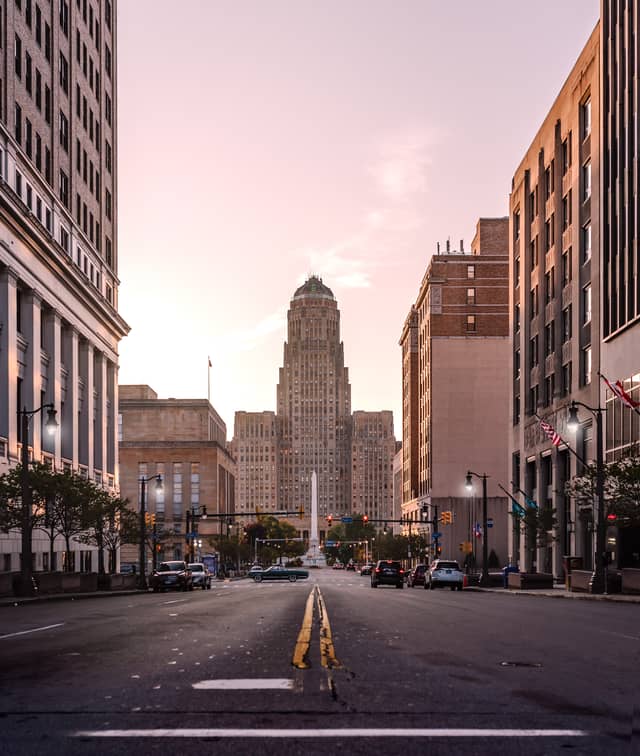 A skyscraper and monument are shown at the end of a road flanked by city buildings