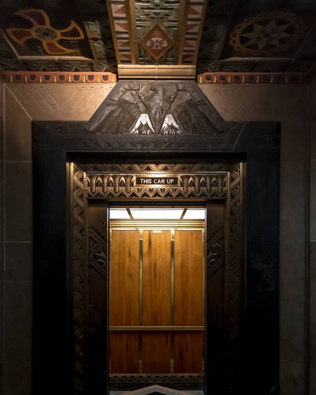 A gold-plated elevator entrance is open with a sign that says 'This car up'