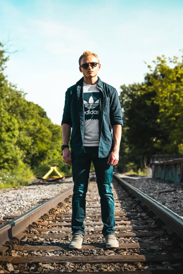 A man with sunglasses is looking head on at the camera while standing on a set of train tracks