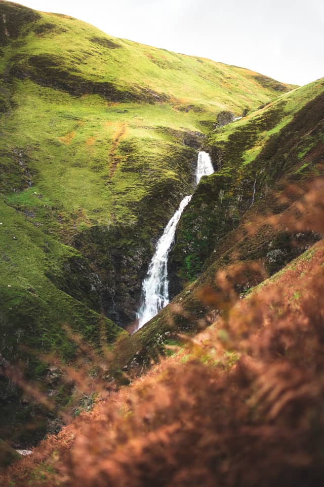 A small waterfall flows over grass-lined hills