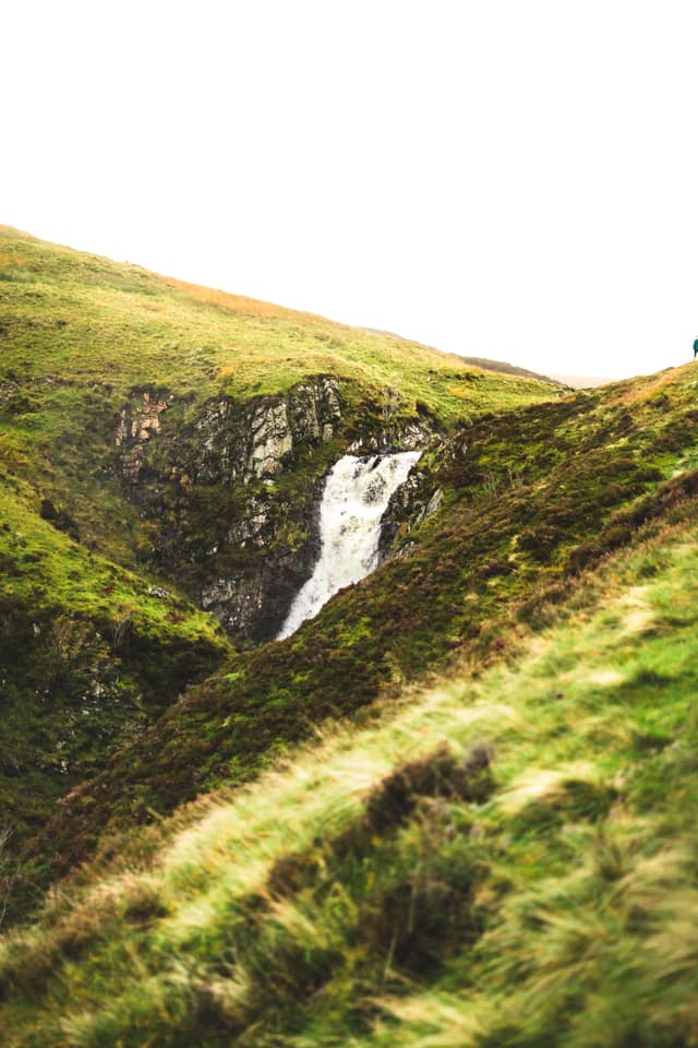 A small waterfall flows over grass-lined hills